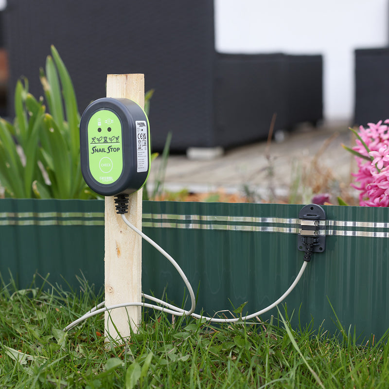 "Kerbl Snail Stop electric fence energizer unit mounted on a wooden stake in a garden, connected to the self-adhesive contact tape on a green slug barrier."